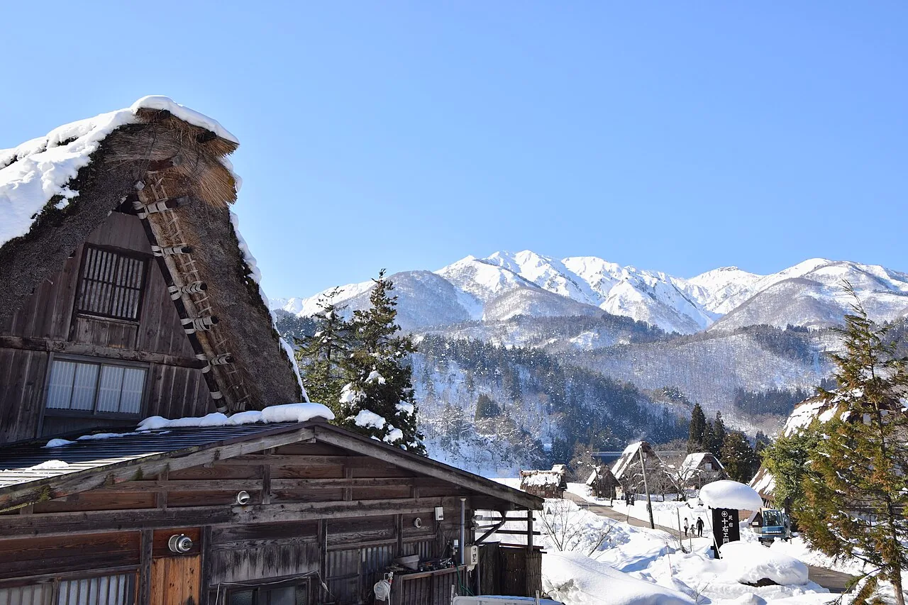 A snow-covered thatched farmhouse in Shirakawa-go with white mountains under a blue sky