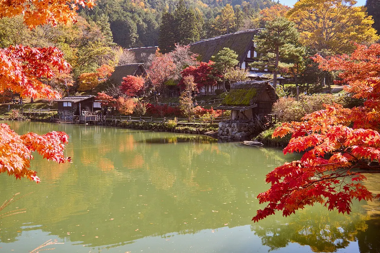 Thatched farmhouses beside a pond framed by red maple leaves at Hida Folk Village in autumn