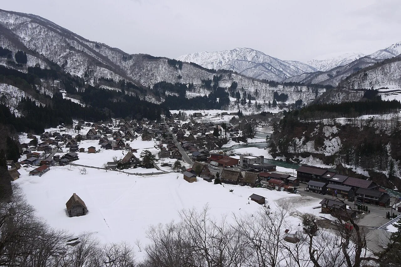 Ogimachi under deep snow on an overcast day, seen from the hillside, with the river and snowy mountains behind
